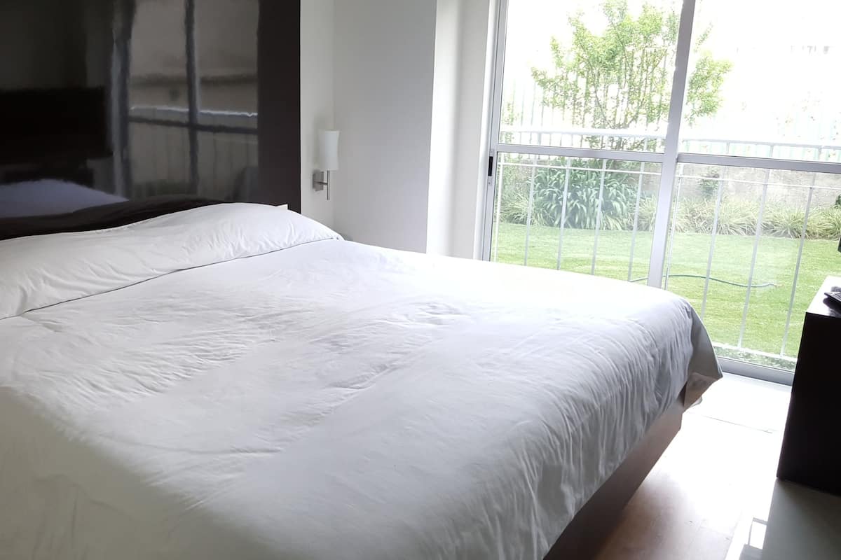 A bedroom with a large bed, a glass table, a TV, and a view of a garden.
