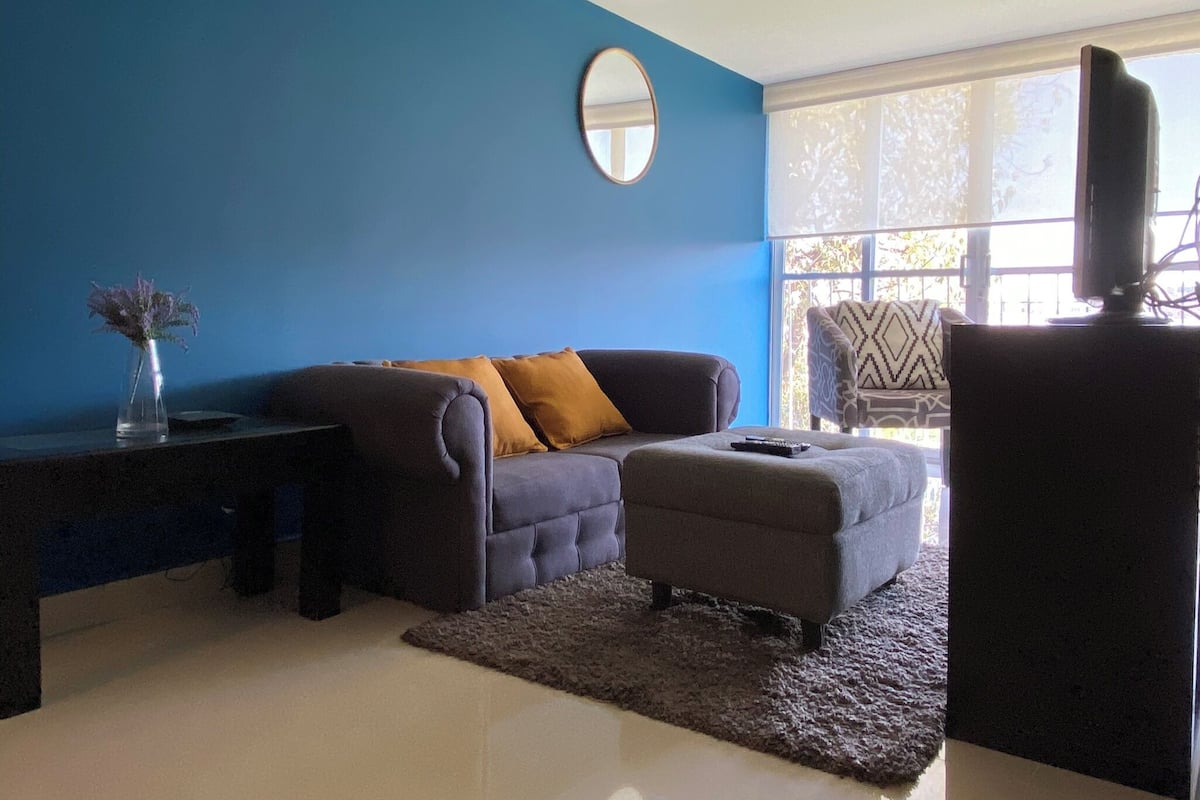 A living room with a blue wall, a round mirror, a grey sofa with a mustard cushion, a small ottoman, and a television on a stand.