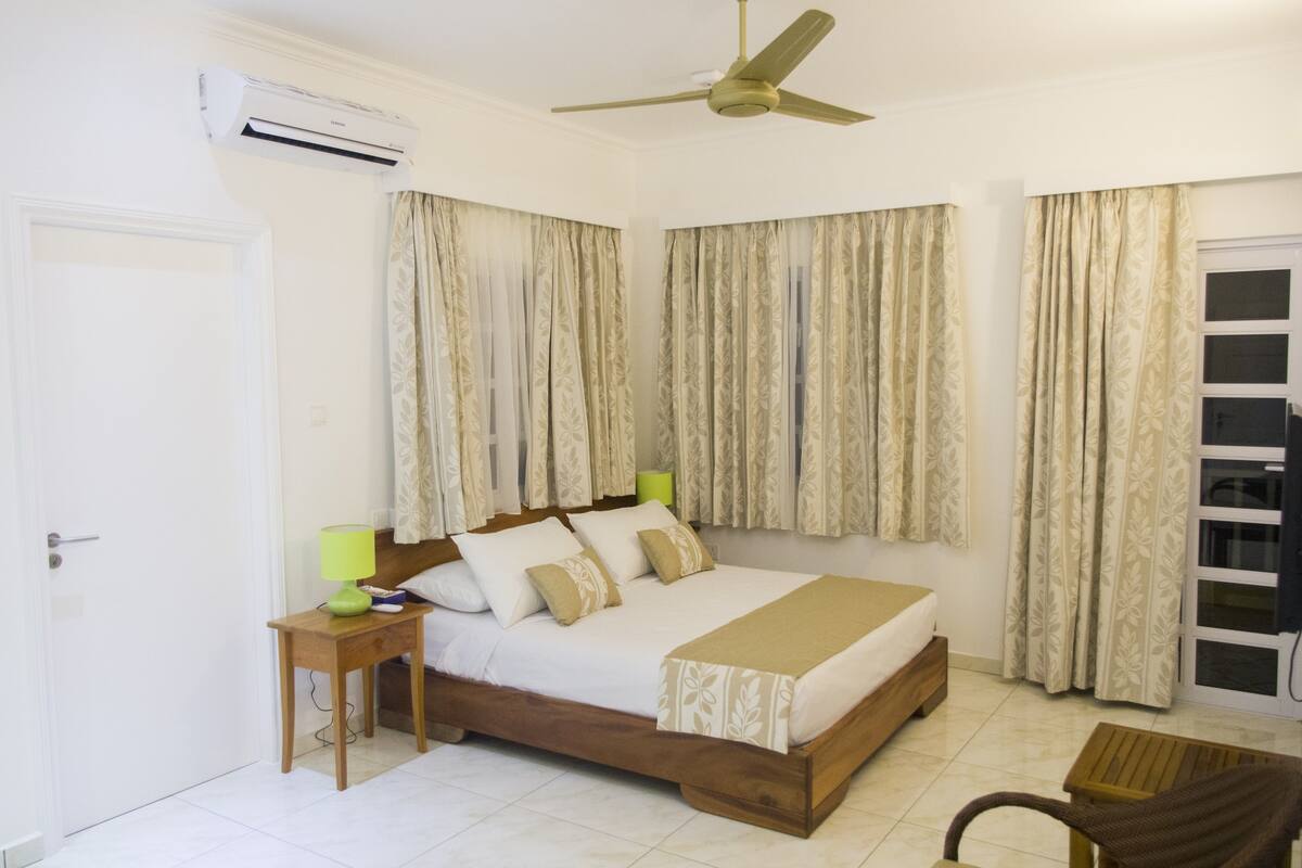 Une chambre à coucher comprenant un lit, une table de chevet, une lampe, un ventilateur de plafond et une fenêtre avec des rideaux.