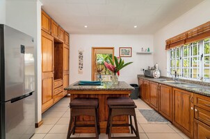 A kitchen with wooden cabinets, a stainless steel refrigerator, a granite countertop, two wicker stools, and a window with blinds.