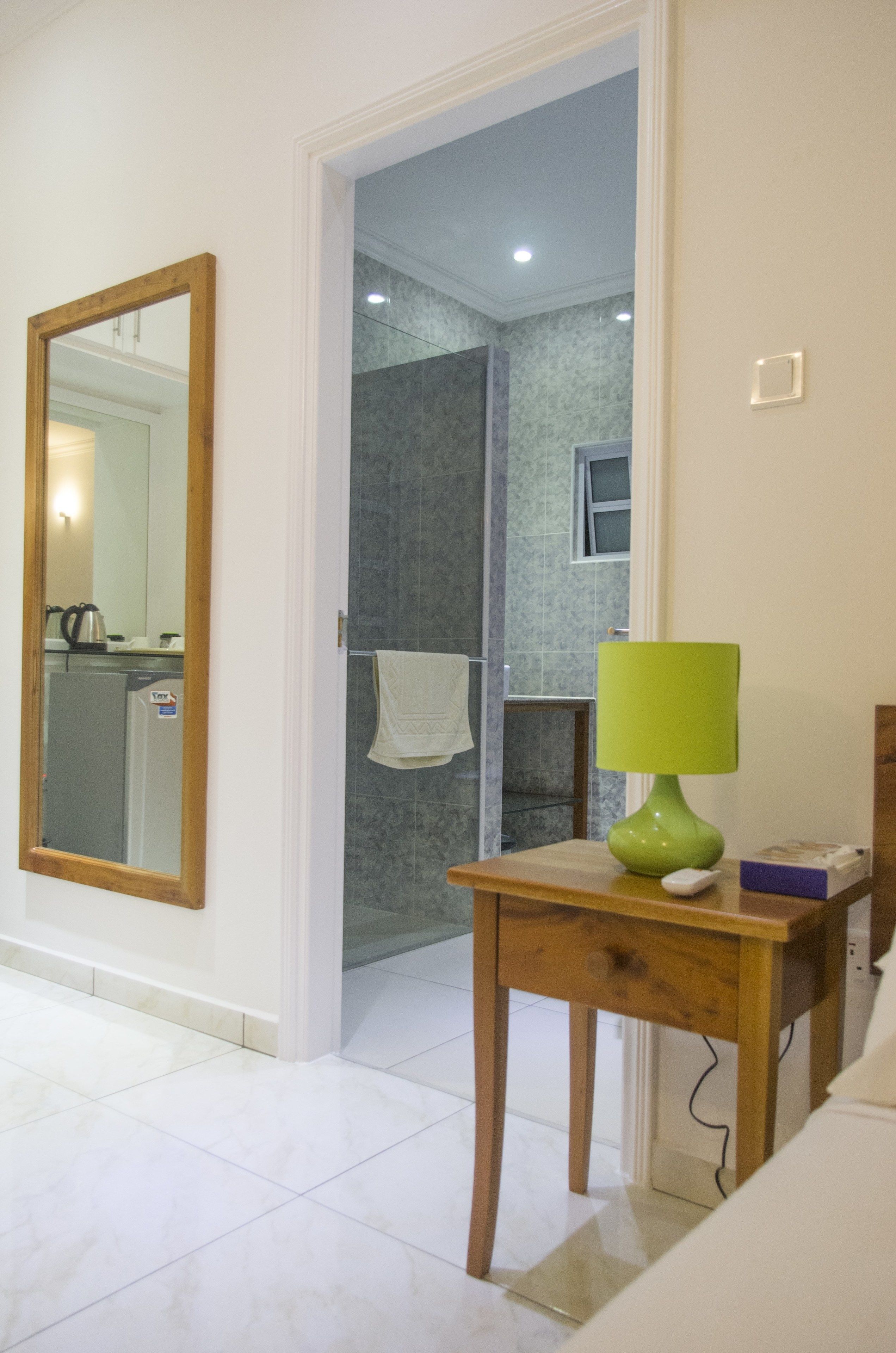 A bathroom with a shower, a mirror, and a green lamp on a wooden table.
