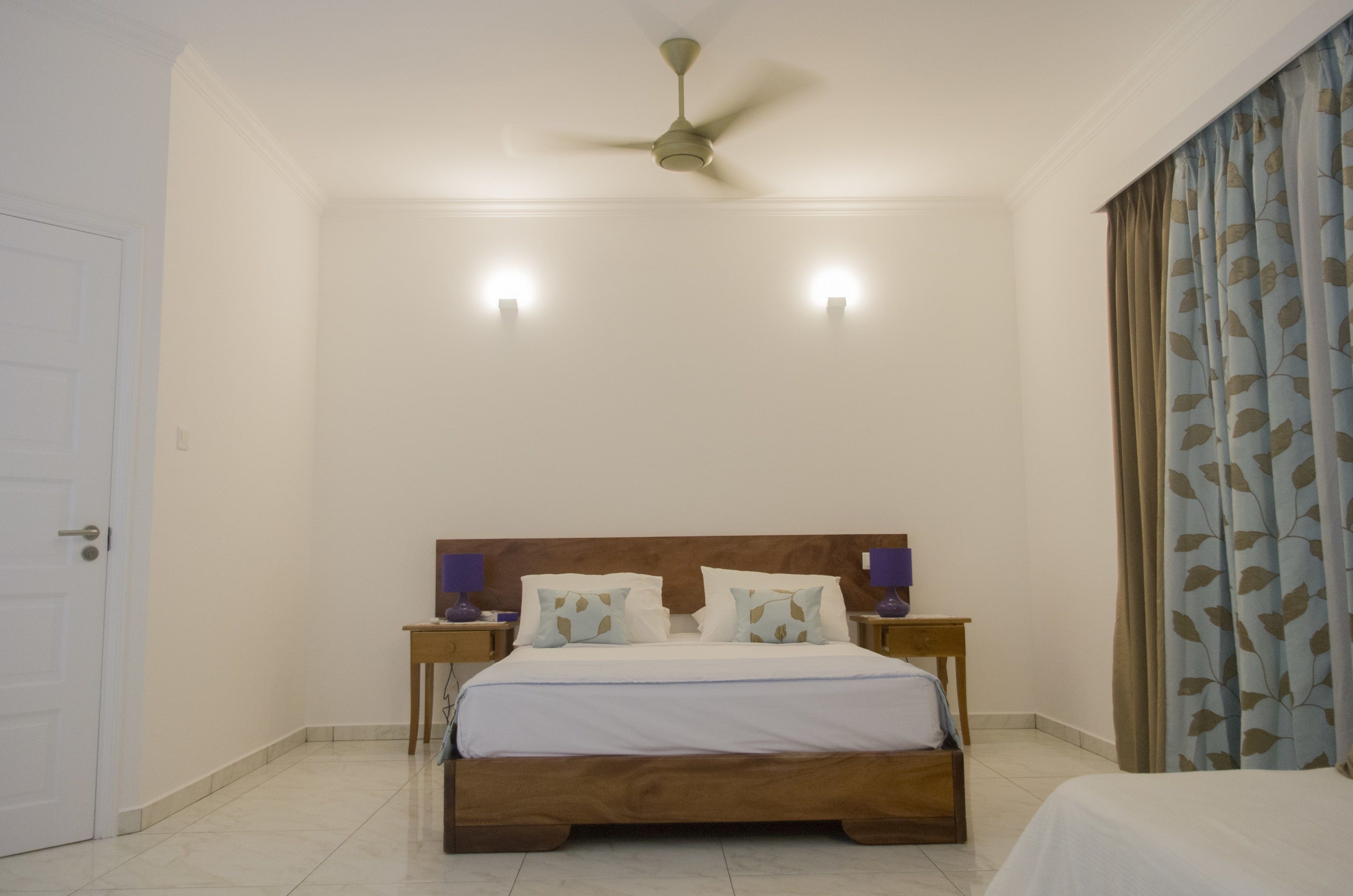A bedroom with a wooden bed, two bedside tables with lamps, a ceiling fan, and a door.