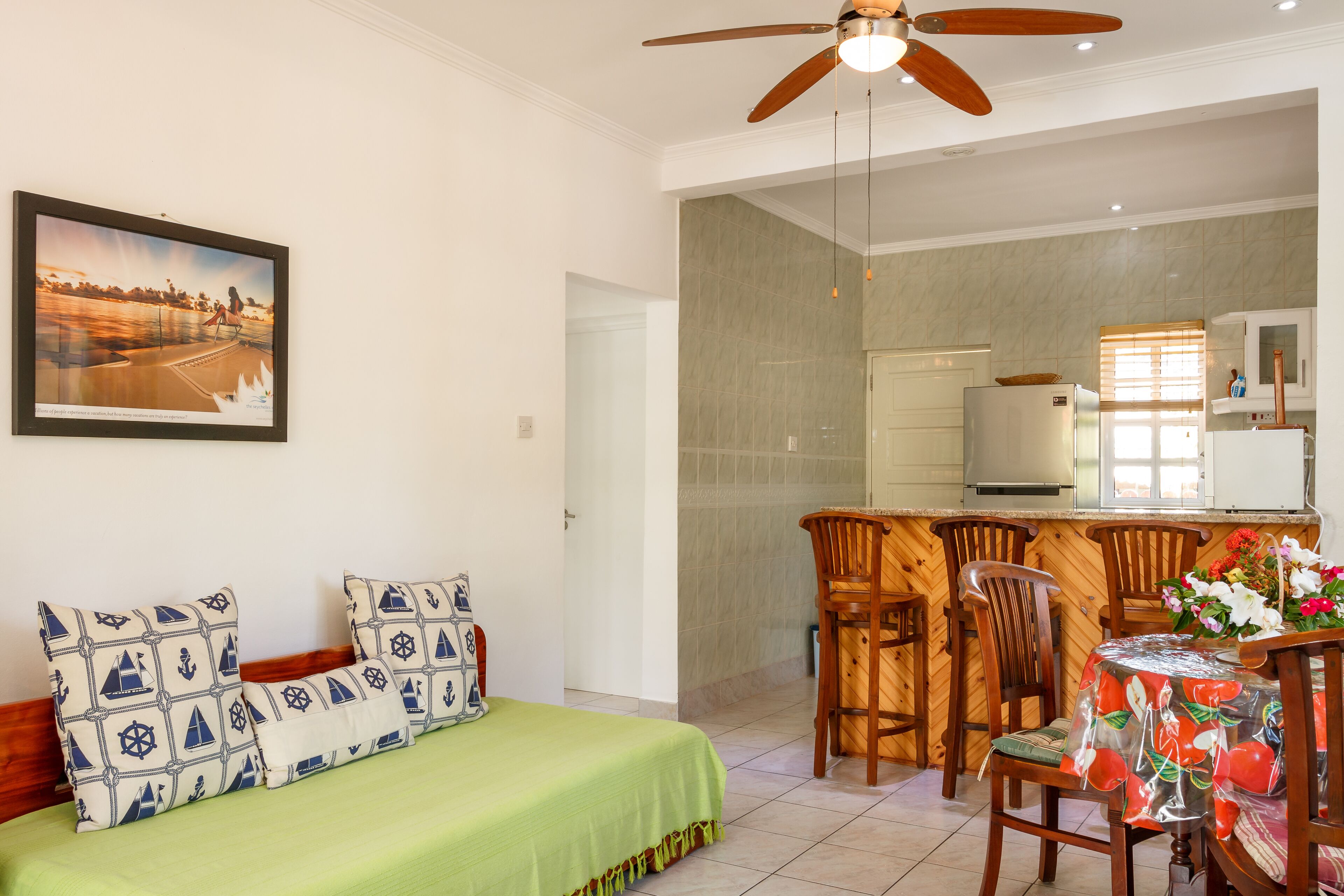 A room with a bed, a dining table with chairs, a kitchen area, and a ceiling fan.