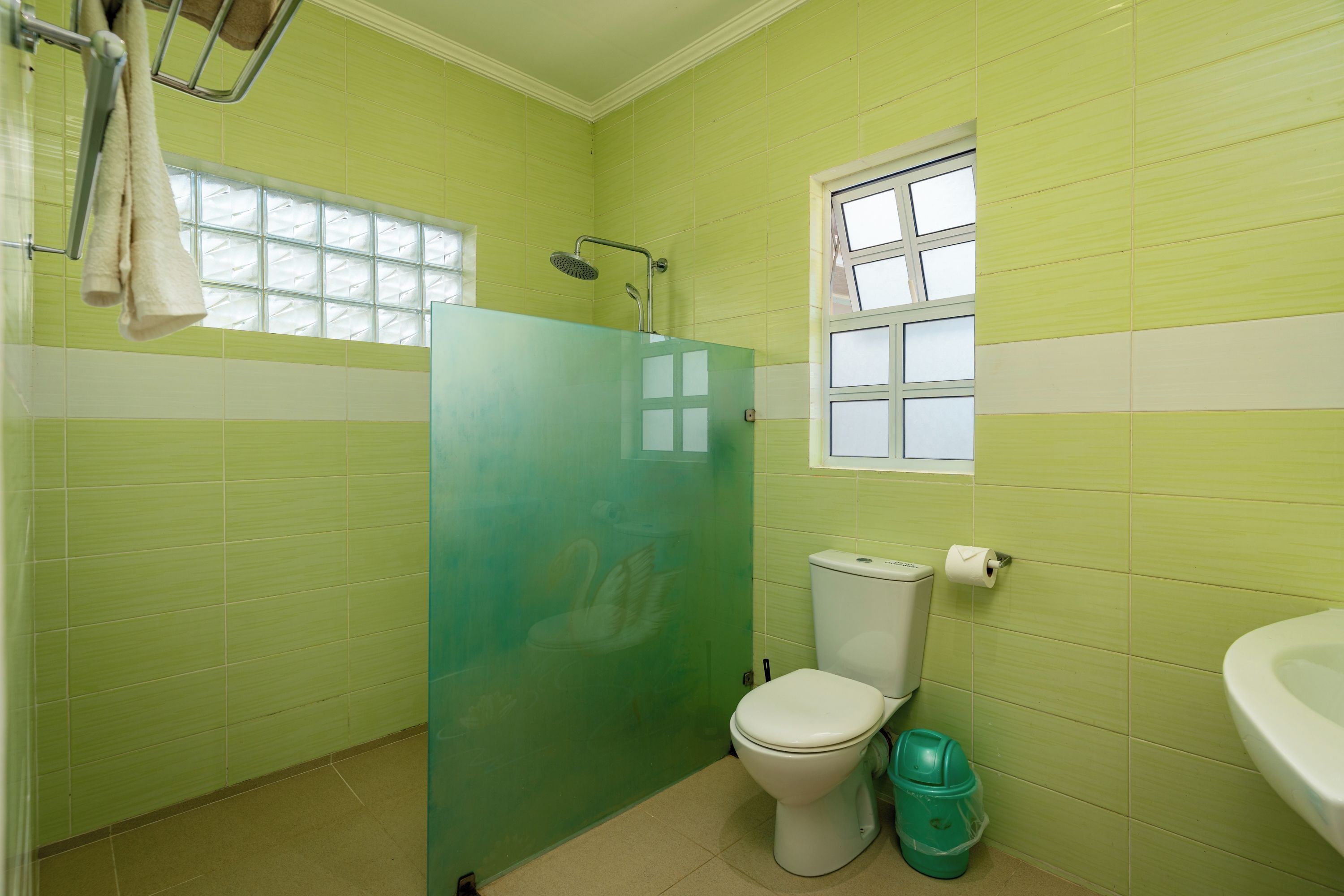 A bathroom with green tiles, a white toilet, a shower, and a window.