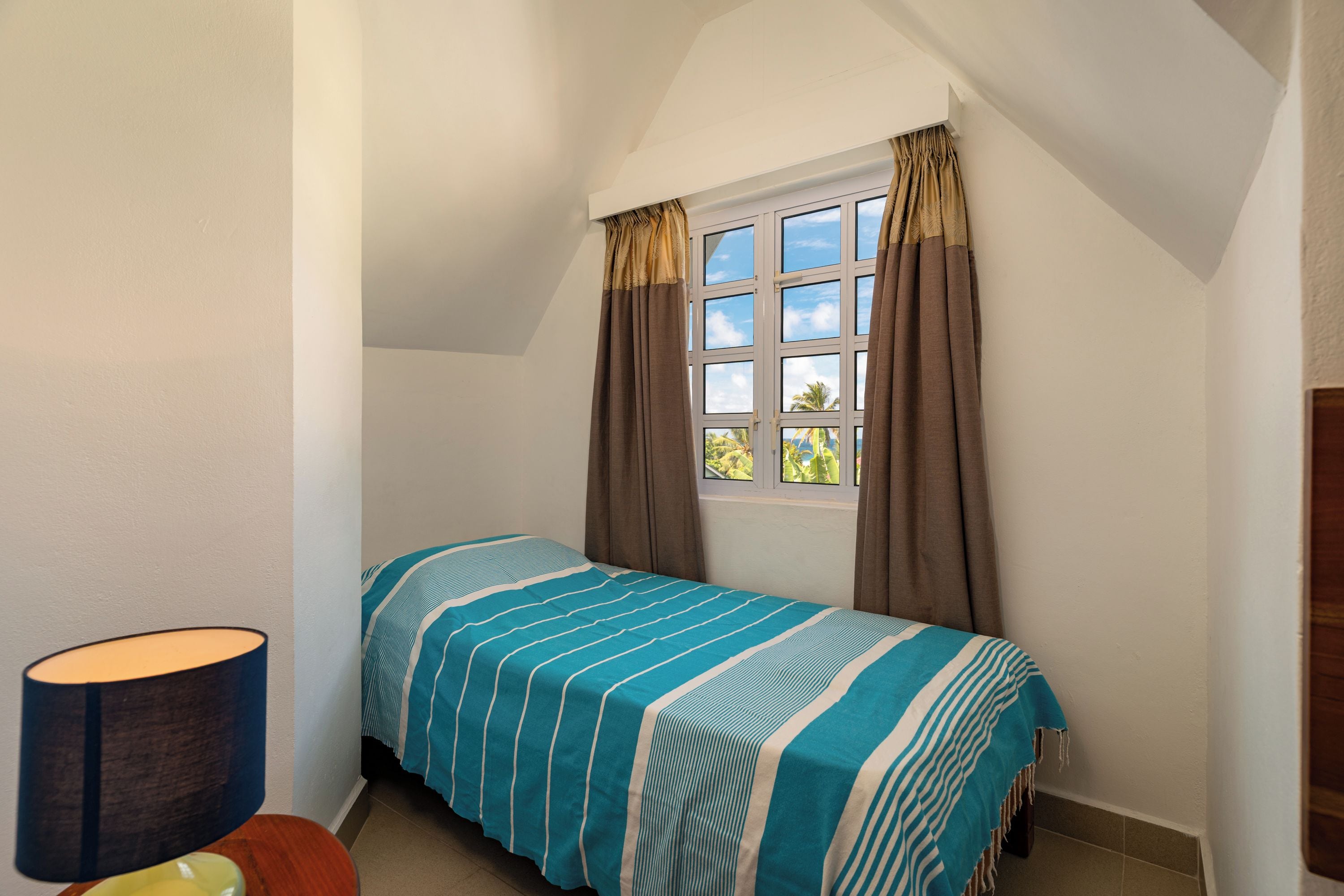 A small, single-bed room with a window, curtains, a bedside lamp, and a view of greenery outside.