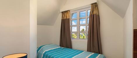 A small, single-bed room with a window, curtains, a bedside lamp, and a view of greenery outside.
