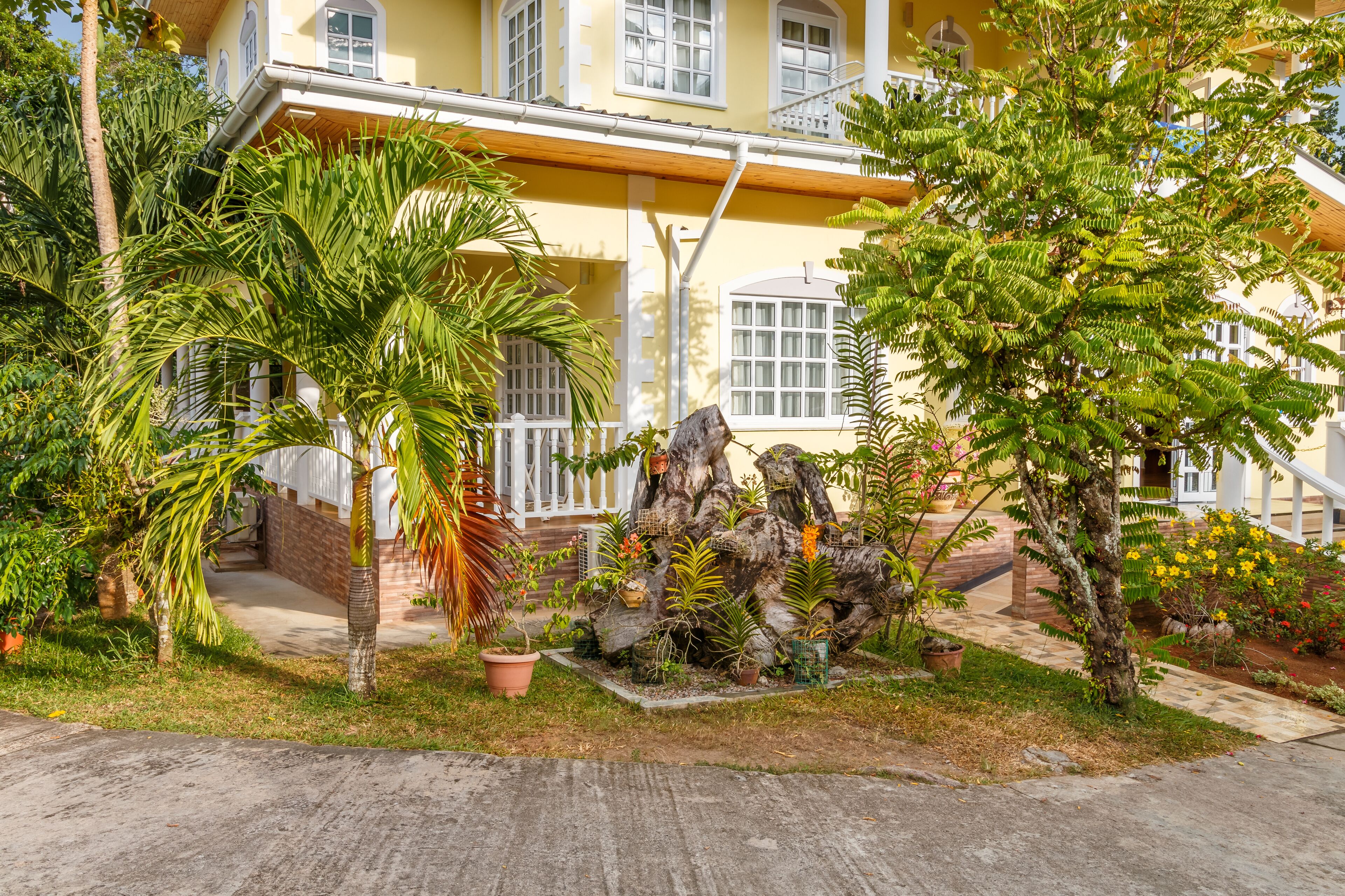 A yellow building with a balcony, surrounded by lush greenery and potted plants.