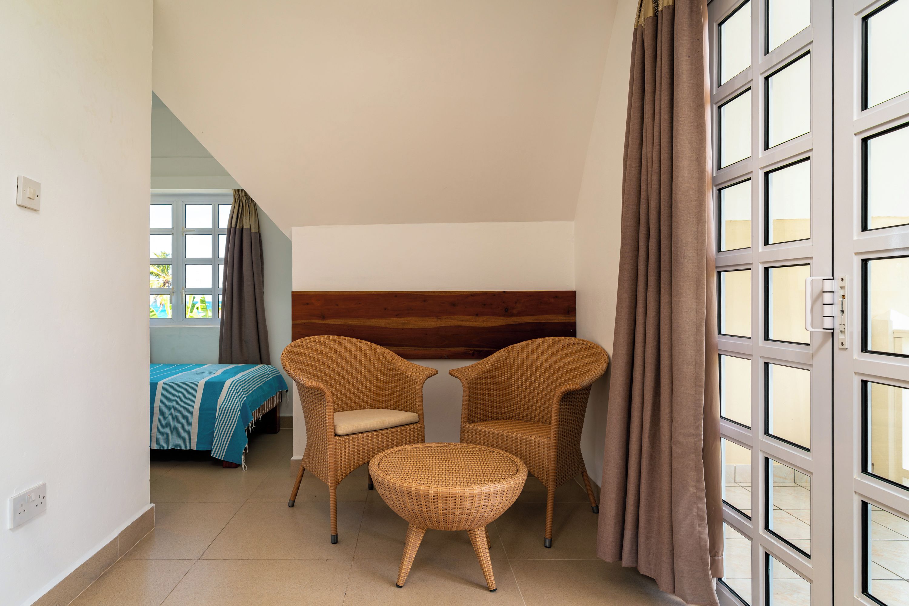 A room with a bed, two wicker chairs, a small table, and a large window with curtains.