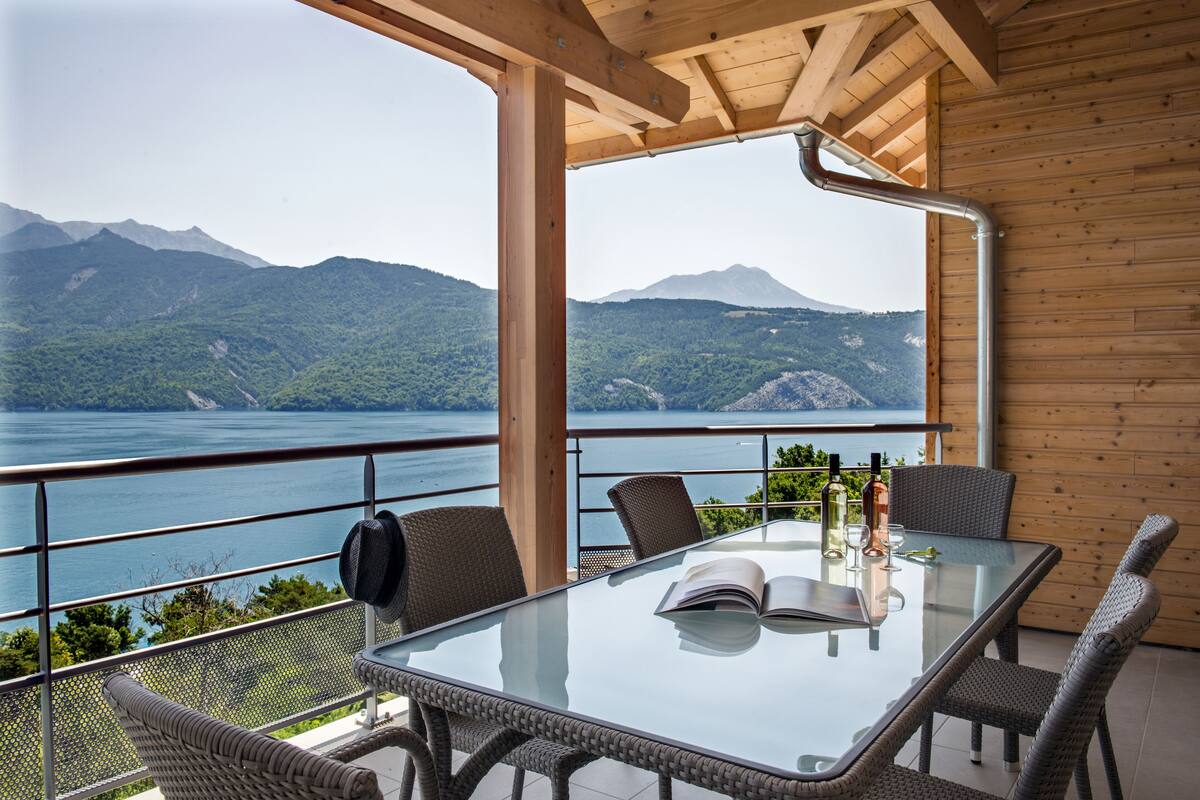 Mesa de vidrio con sillas de mimbre, un libro y botellas en un balcón con vistas a un lago y montañas.