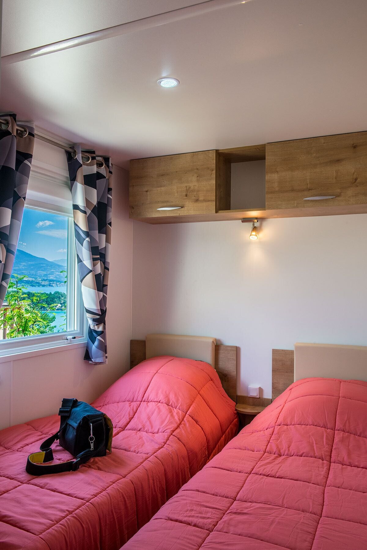 A bedroom with two beds, a window with curtains, a wooden shelf, and a wall-mounted light.