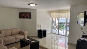 A modern living room with a sofa, a coffee table, a side table with a lamp, a TV, and a large window with a view of the outdoors. - Grupo Kings Suites Chimborazo (Mexico City)
