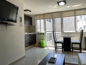 A modern living room with a flat-screen TV, a dining table with chairs, a sofa, and a large window with a view.