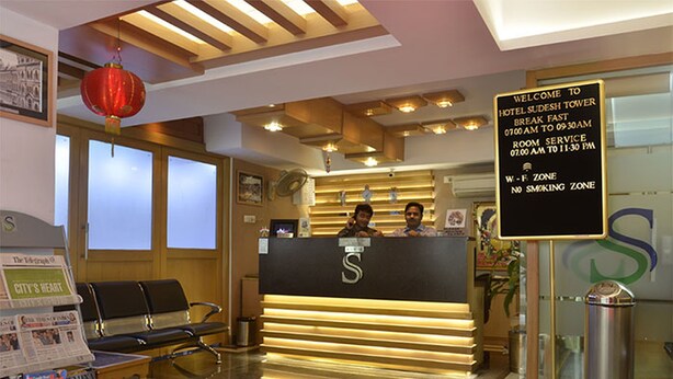 A hotel reception area with a reception desk, two staff members, a 'W-F-ZONE' sign, and a 'CITY'S HEART' newspaper rack. - Hotel Sudesh Tower (Kolkata)