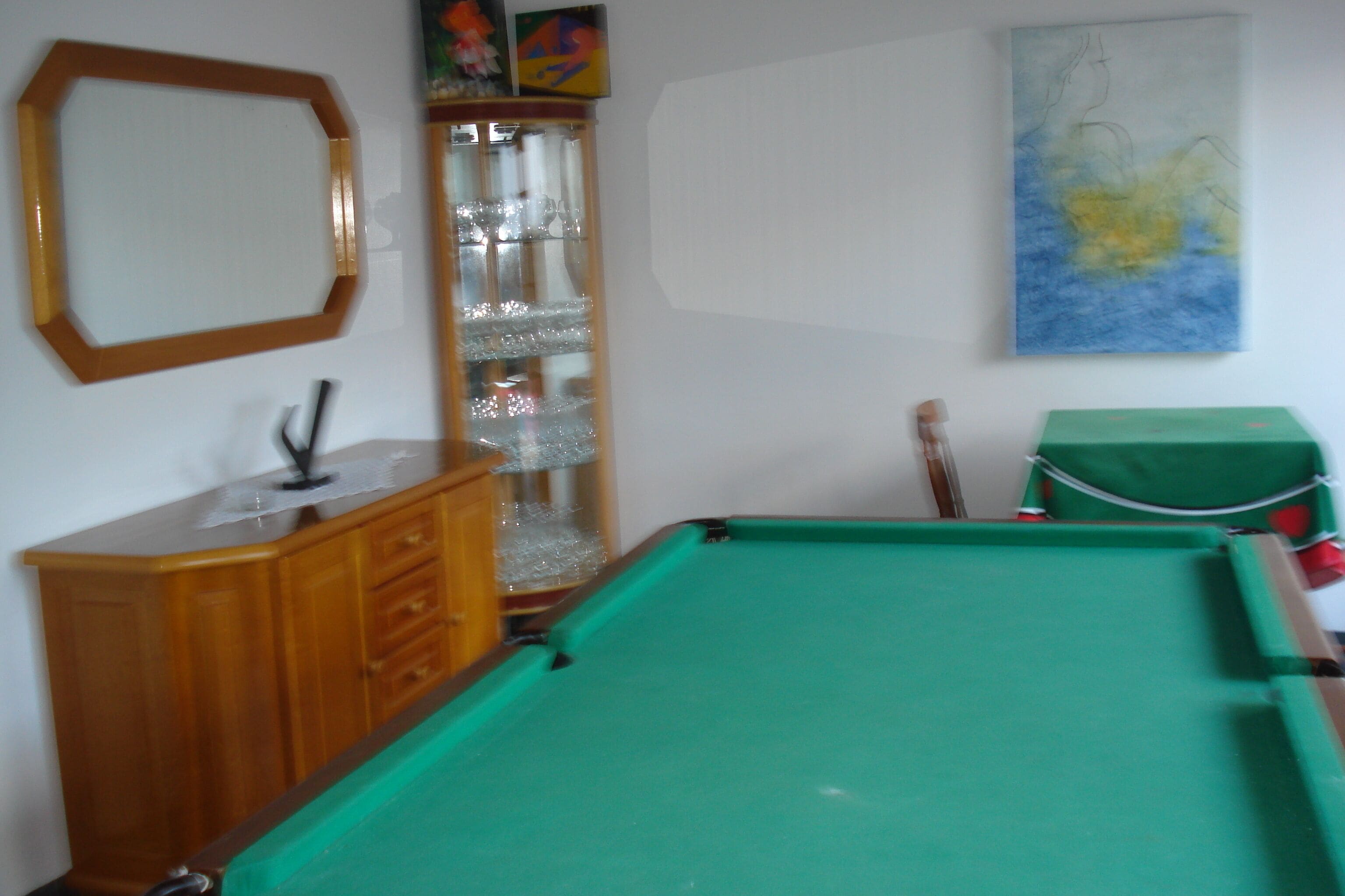 Een pooltafel met een groen vilt oppervlak, een houten kast met lades, een glazen displaykast met decoratieve objecten, en een schilderij aan de muur.