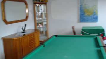 Een pooltafel met een groen vilt oppervlak, een houten kast met lades, een glazen displaykast met decoratieve objecten, en een schilderij aan de muur.