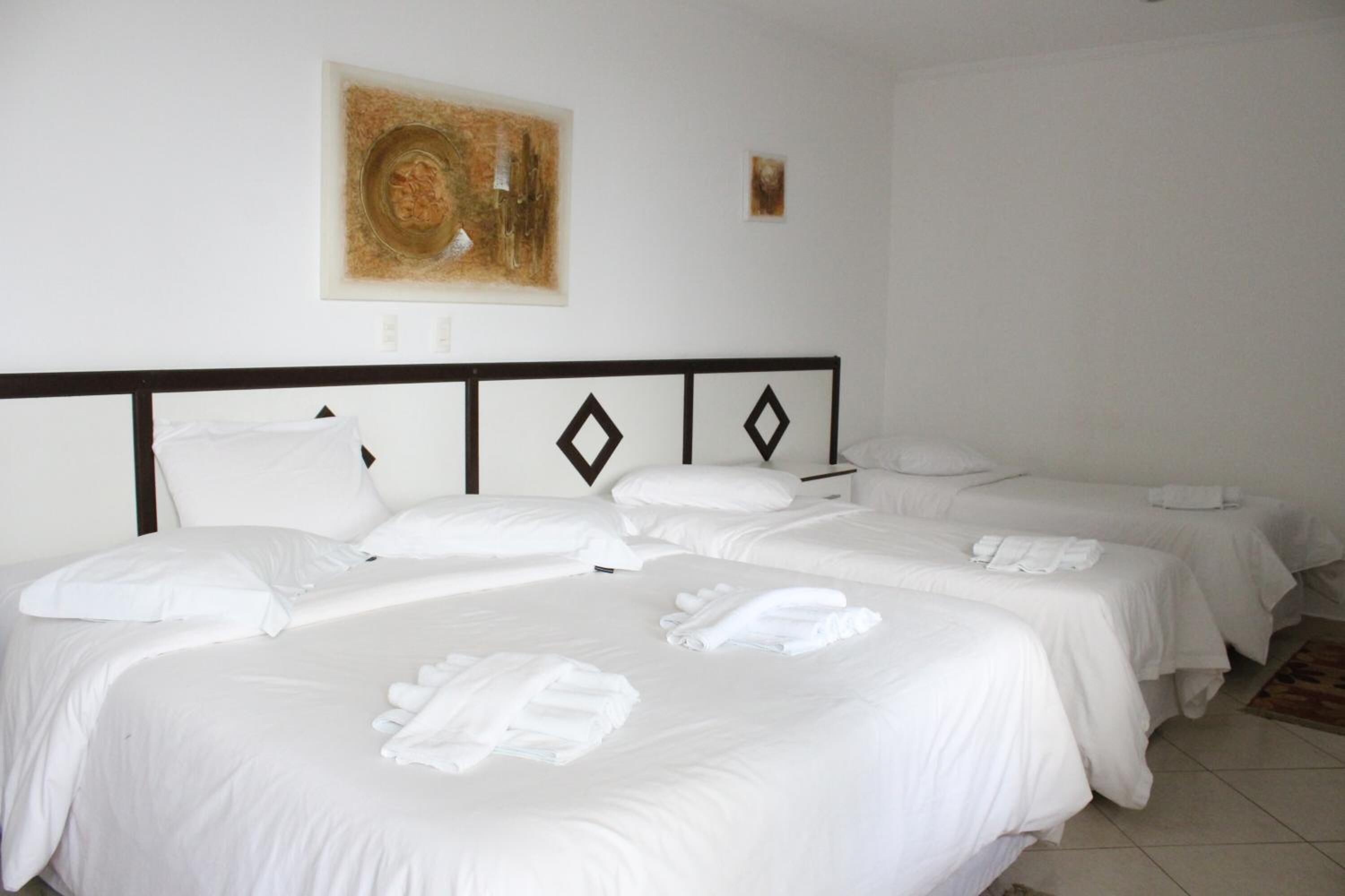 Una habitación de hotel con una cama grande, dos camas más pequeñas, ropa de cama blanca, un cabecero con diseños de diamantes y un cuadro en la pared.