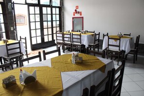 A dining area with multiple tables, each covered with a yellow tablecloth and white tablecloths. There are wooden chairs around the tables. A fire alarm is mounted on the wall. - Hotel Pousada da Néia (Cananeia)