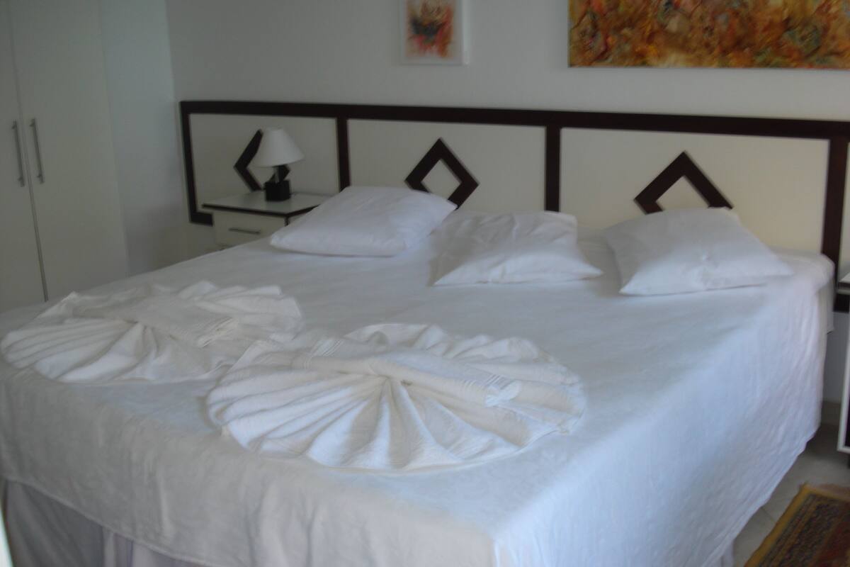 A neatly made bed with pillows and a folded towel in the center, a bedside table with a lamp, and a painting on the wall.
