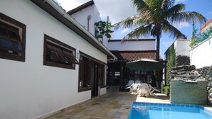 Un área de piscina con suelo de baldosas, un edificio blanco con marcos de ventanas oscuros y una palmera.