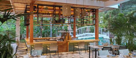 Un área de comedor al aire libre con mesas y sillas, un bar, y vistas a una piscina y follaje.