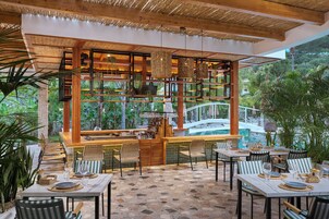 Uma área de refeições ao ar livre com mesas e cadeiras, um bar e vista para uma piscina e vegetação.