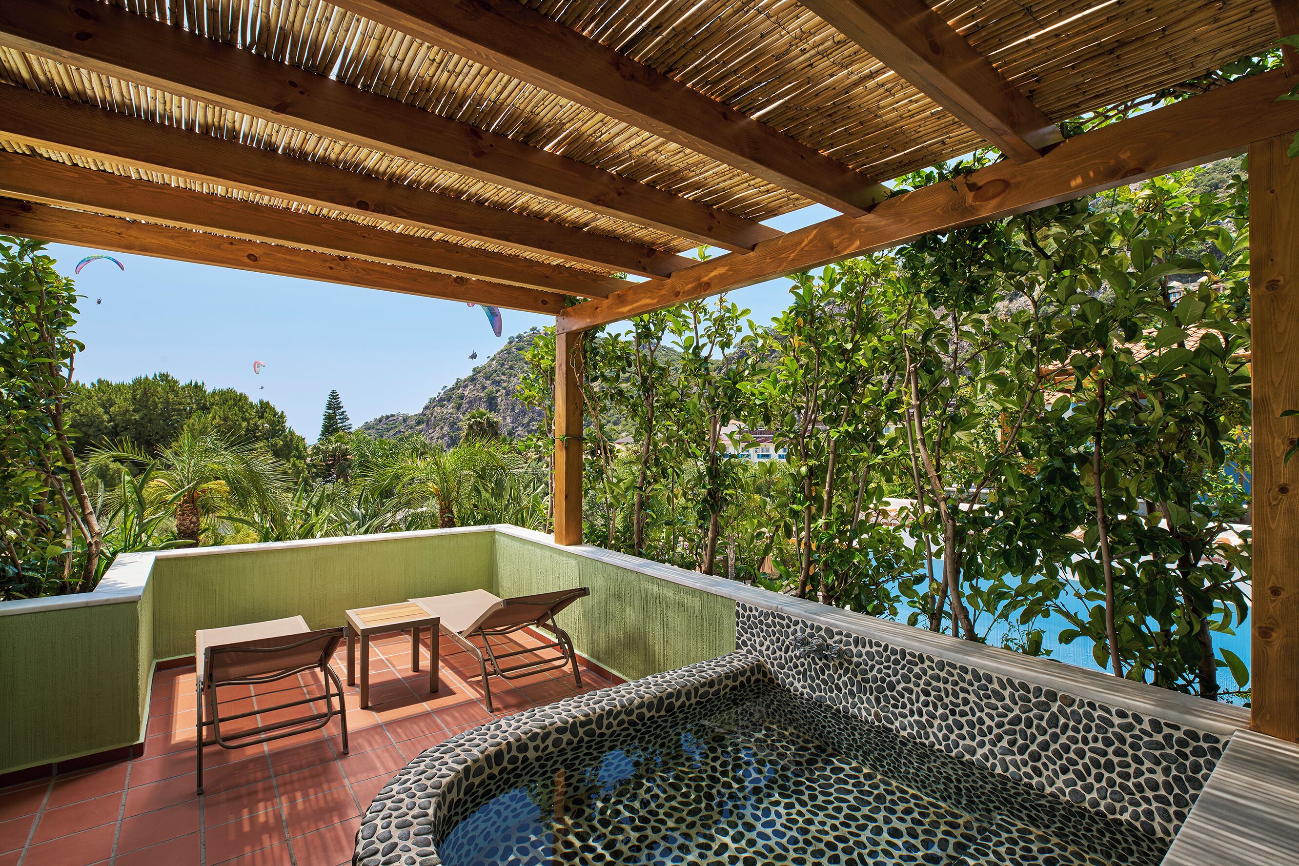 Una terraza en la azotea con un jacuzzi de piedra, mobiliario de madera y vistas a la vegetación exuberante y montañas.