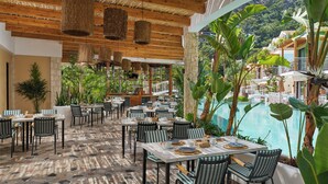Un área de comedor al aire libre con mesas y sillas, rodeada de abundante vegetación y una piscina.