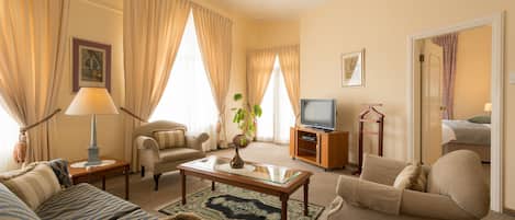 Habitación de hotel con sofá, sillón, una mesita con lámpara, televisor sobre un mueble de madera y un ventanal con cortinas.
