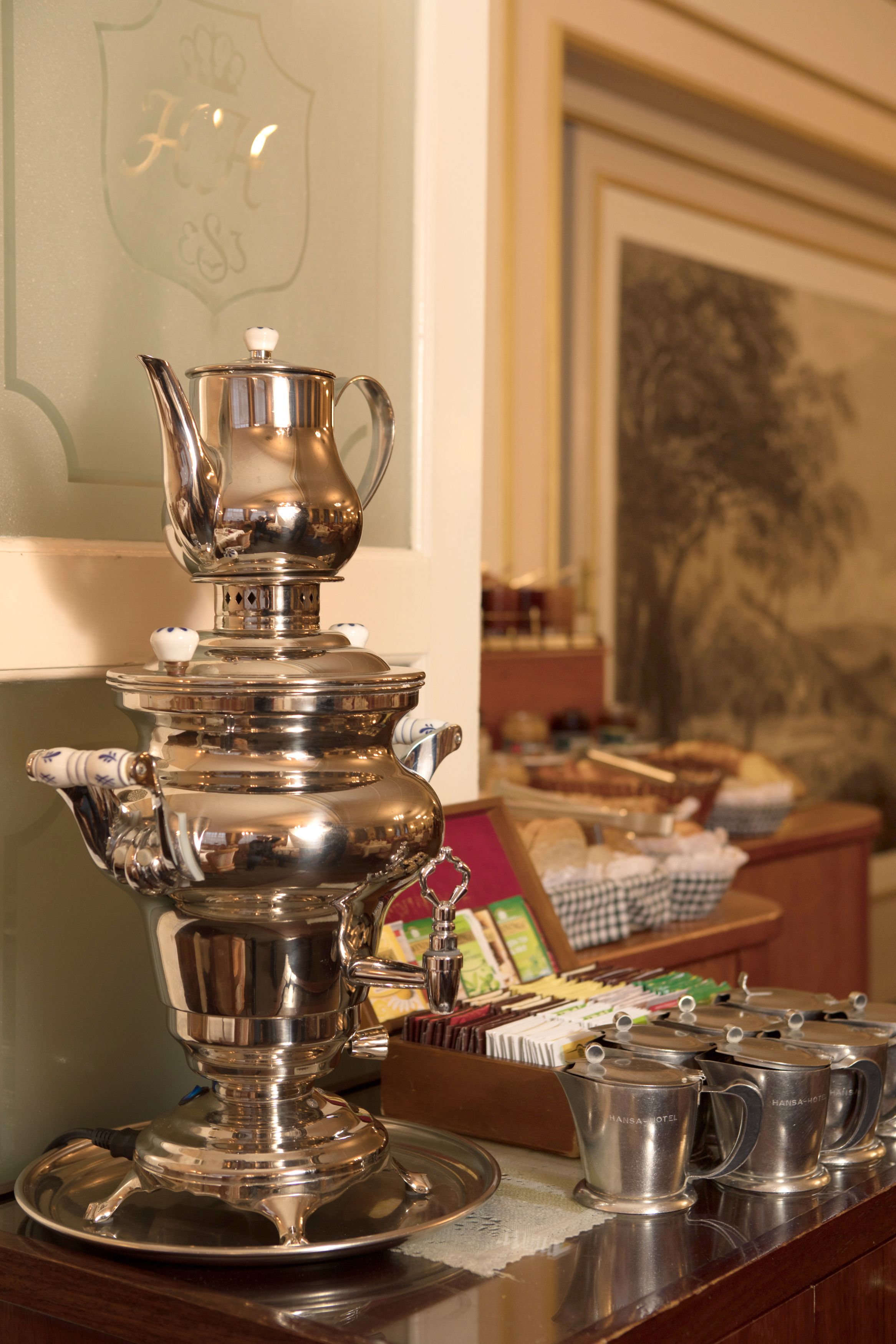 A silver tea service set on a wooden table with a framed picture and a basket of pastries in the background.
