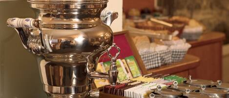 A silver tea service set on a wooden table with a framed picture and a basket of pastries in the background.