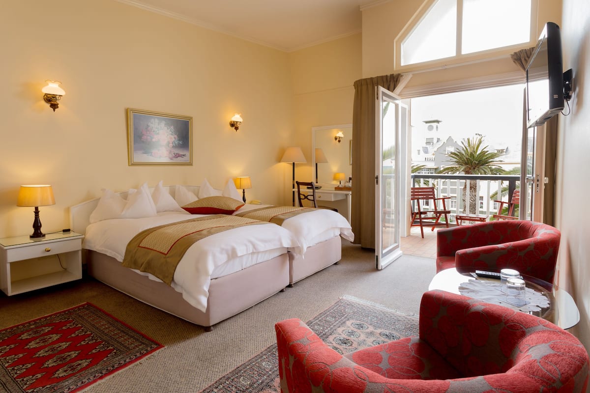 Une chambre d’hôtel avec un lit, deux tables de chevet avec des lampes, un fauteuil rouge, une petite table avec une télécommande et un balcon avec des chaises et une plante.