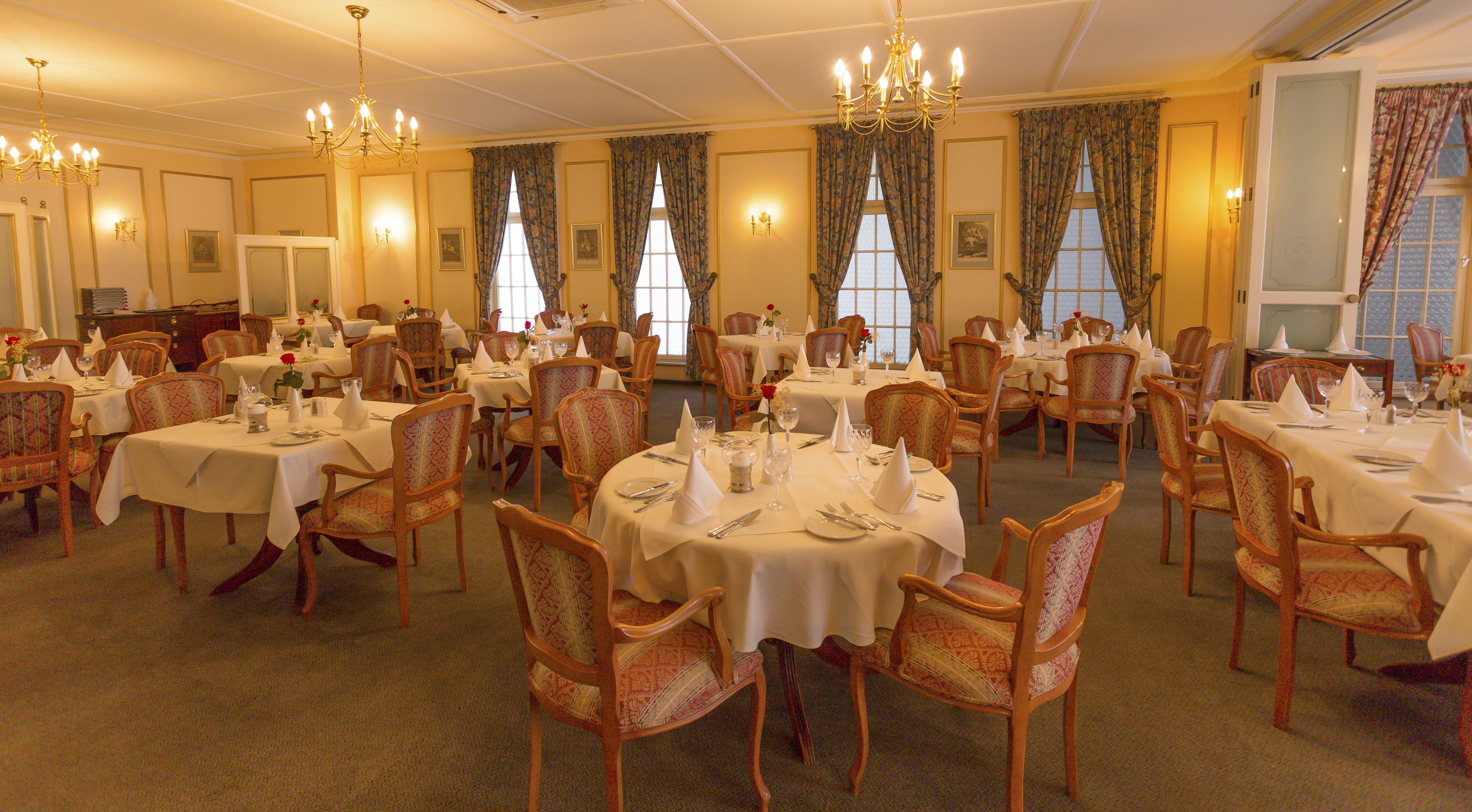 Une salle à manger élégante, avec plusieurs tables rondes dressées pour le service, des chaises en bois recouvertes d’un tissu à motifs et de grandes fenêtres ornées de rideaux à fleurs.