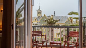 A balcony with two wooden chairs, a view of a building with a clock tower, and palm trees. - Hansa Hotel (Swakopmund)