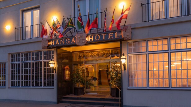 The exterior of Hansa Hotel with multiple flags, large windows, and a staircase leading to the entrance. - Hansa Hotel (Swakopmund)