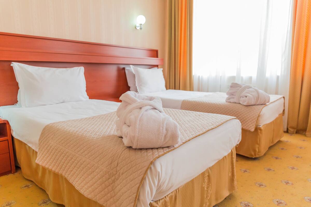 A hotel room with two beds, each with a white comforter and a beige quilted bedspread. There are two white pillows on each bed, and a rolled towel on the beds. The room has a wooden headboard, a nightstand with a lamp, and a window with curtains.