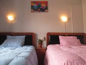 Dos camas dobles con ropa de cama de colores diferentes, una mesita con una planta y un cuadro llamativo en la pared.