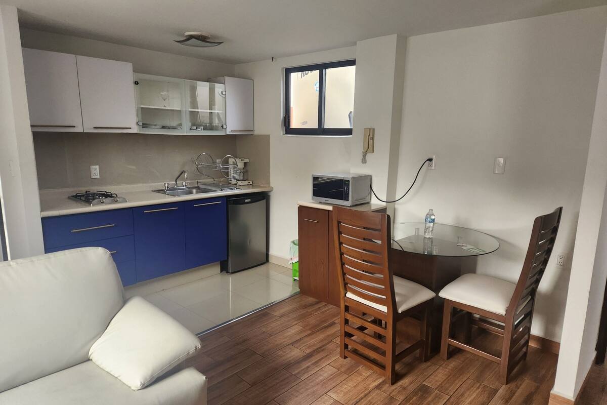 Una cocina compacta equipada con lavavajillas, microondas y fregadero. Cuenta con una zona de comedor con una mesa de vidrio y sillas de madera.