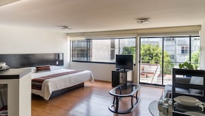 A modern hotel room with a large bed, a TV, a glass table, and a balcony with a view of buildings and greenery. - Grupo Kings Suites Platon 436 (Mexico City)