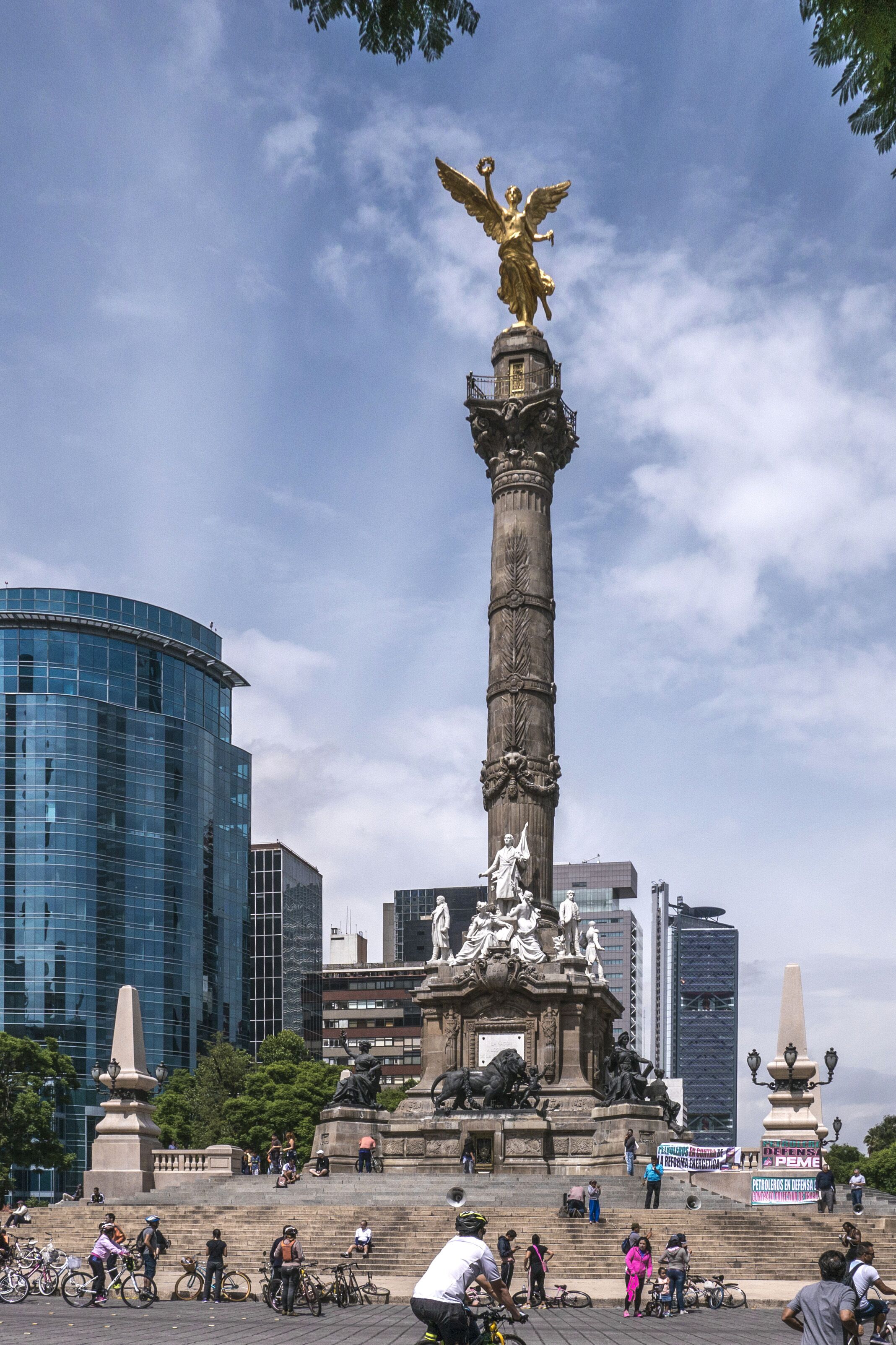 A tall monument with a golden angel statue, surrounded by a plaza with steps and people.