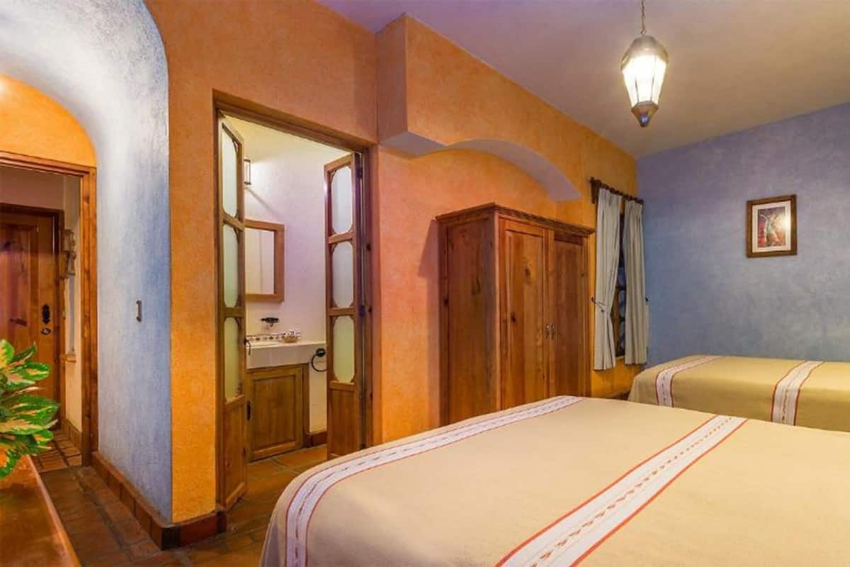 A hotel room with two beds, a wooden wardrobe, a bathroom with a mirror, and a hanging light fixture.
