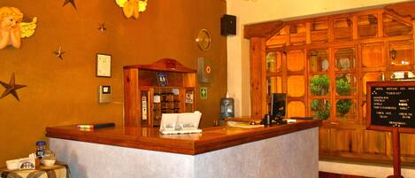 A hotel reception area with a wooden reception desk, a clock, and a signboard with room rates.