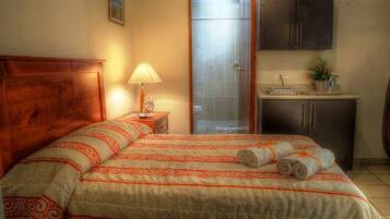 Habitación de hotel con cama, mesita de noche con lámpara, baño con ducha y zona de kitchenette.