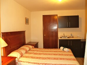 A bedroom with a wooden bed, a nightstand, a bedside lamp, a door, and a kitchen area with a sink and cabinets. - Hotel & Posada Refugio Independencia (Morelia)