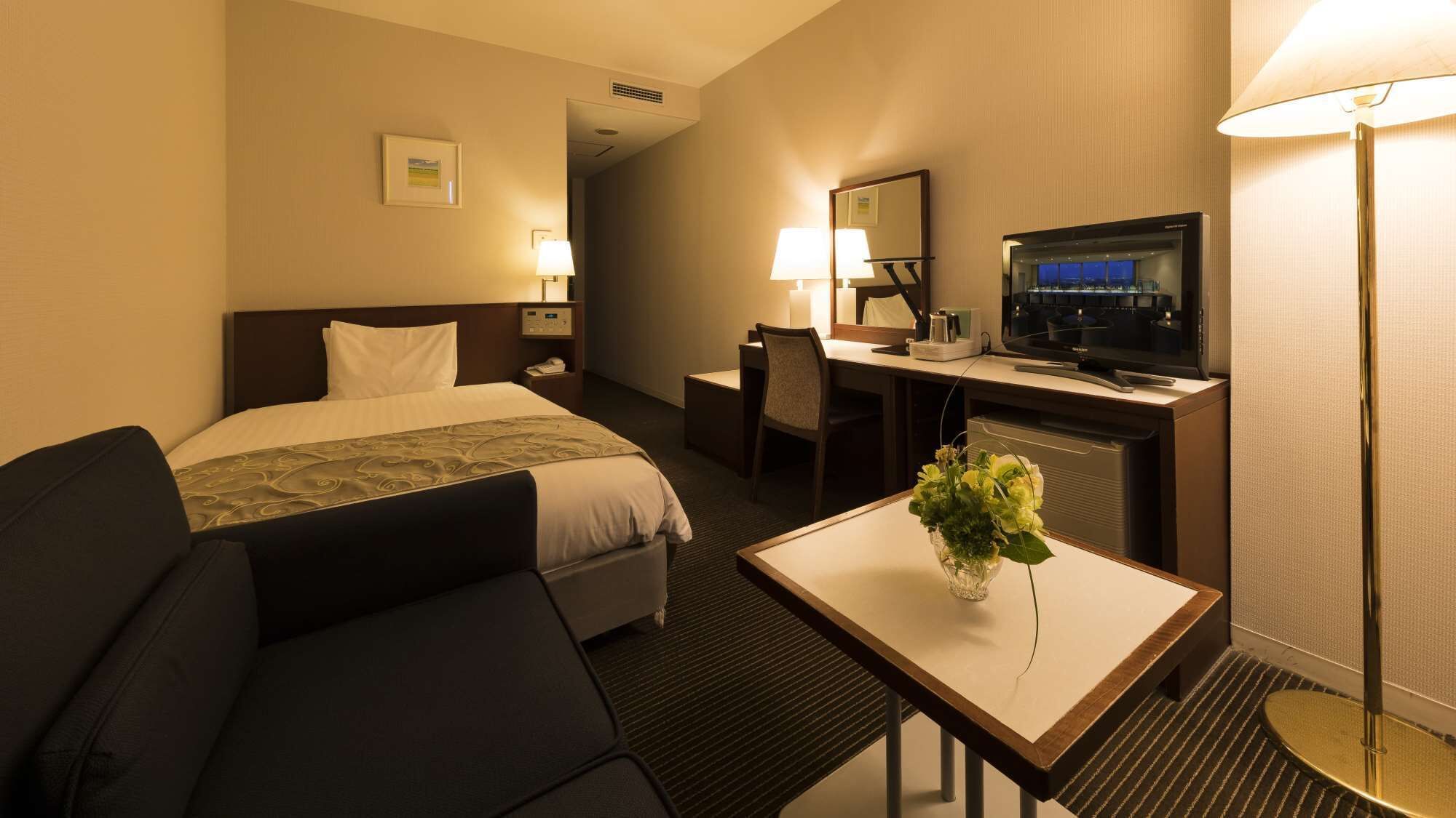 A hotel room with a bed, a desk with a TV, a chair, a small table with a vase, and a lamp.