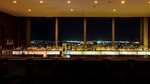 Un bar con vistas a la ciudad de noche, que presenta una larga barra con diferentes botellas y vasos, y varias zonas de estar al frente.