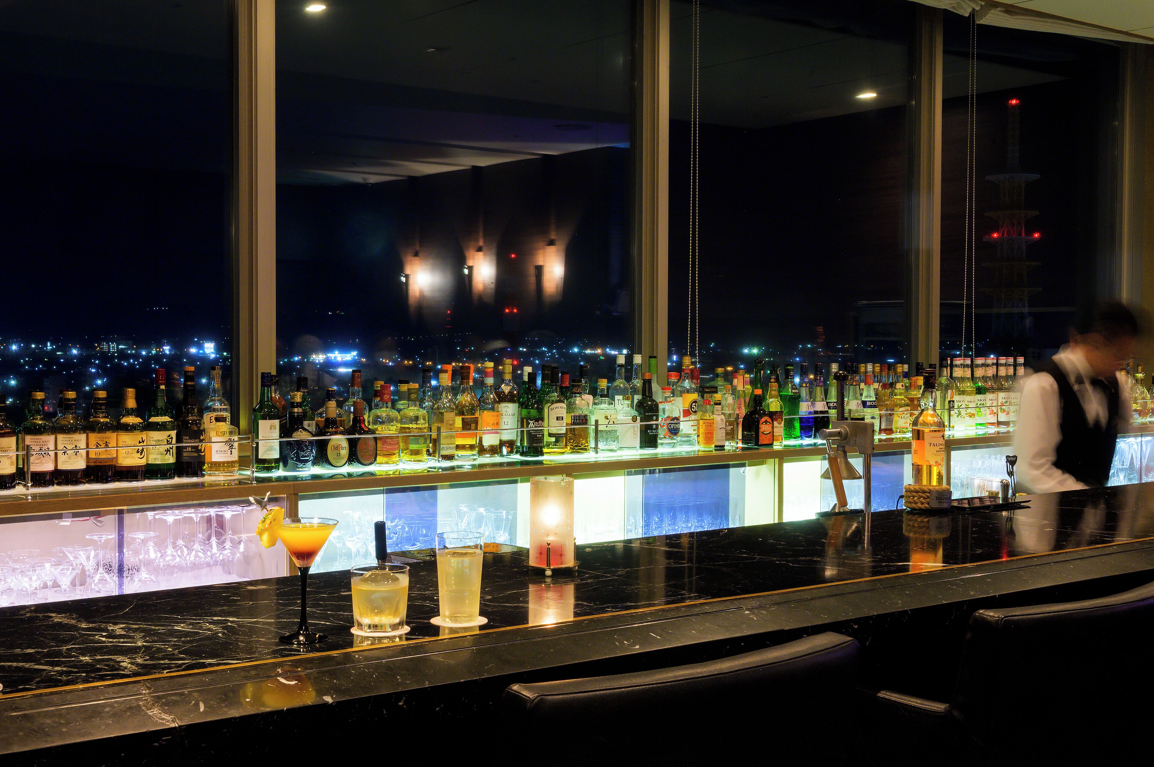 A bar with a city view at night, bottles of alcohol, and a bartender.