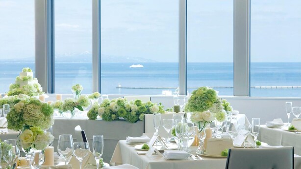 Un comedor con mesas dispuestas para un evento formal, amplios ventanales con vistas al mar y un barco a lo lejos.