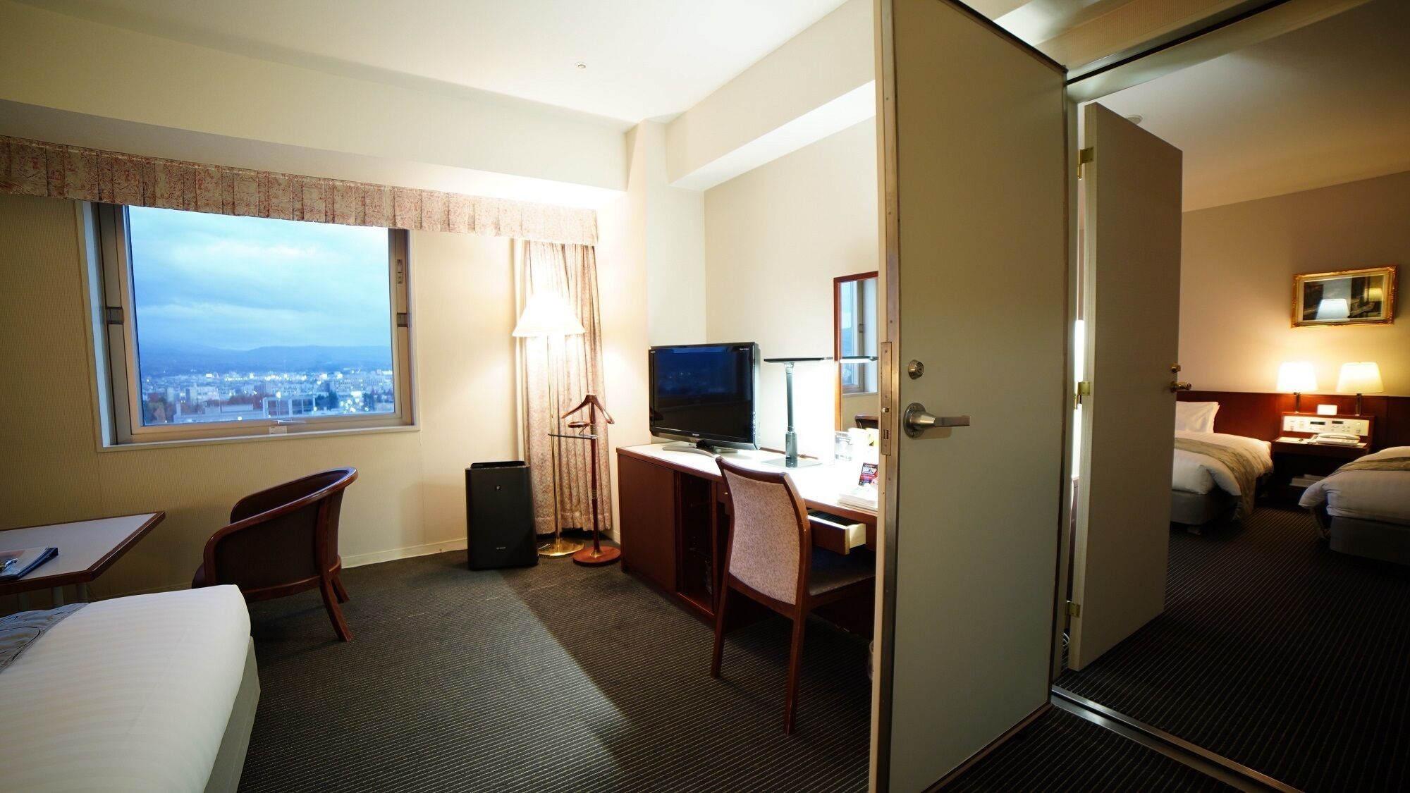 A hotel room with a bed, desk, chair, TV, and a city view.