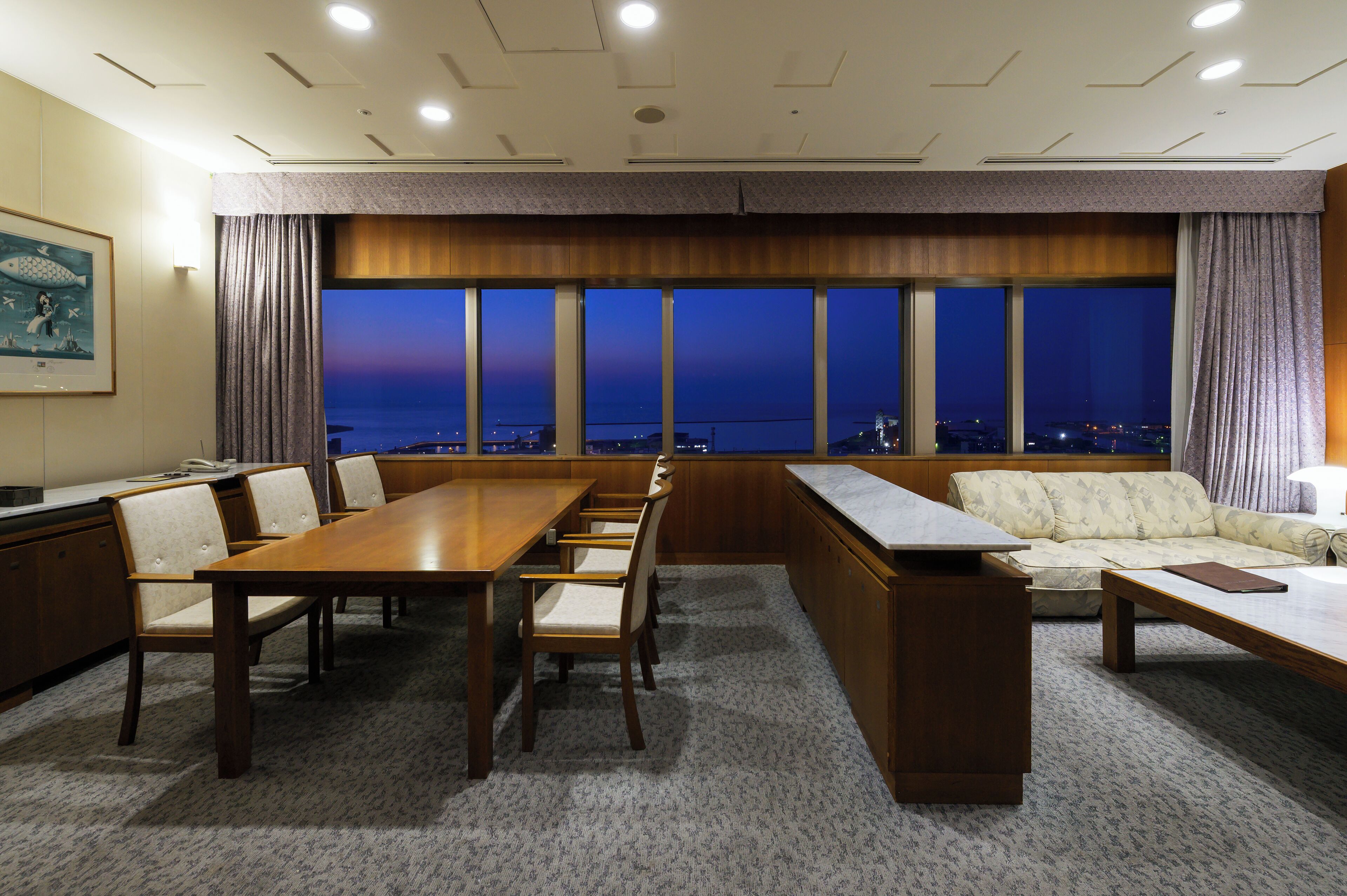 Una sala de conferencias con una larga mesa, sillas, un sofá y un amplio ventanal con vista a la ciudad al atardecer.
