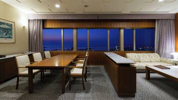 Una sala de conferencias con una larga mesa, sillas, un sofá y un amplio ventanal con vista a la ciudad al atardecer.